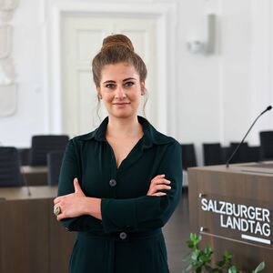 Portraitbild von Natalie Hangöbl, Klubobfrau der KPÖ Plus im Salzburger Landtag, mit verschränkten Armen und lächelnd vor dem Rednerpodium des Salzburger Landtags.