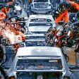 Mehrere große Roboterarme arbeiten an einer Reihe silbergrauer Autos im Rohbau, Funken sprühen.