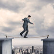 A businessman balancing on a tightrope high above a big city. He looks down at the rope before his next step as he nears the midpoint of the tightrope. Metal spikes are anchored into the concrete to help support the tightrope. The New York City skyline can be seen in the distance as a fog persists over the buildings.