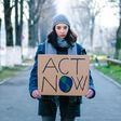 Portrait of a young teenage girl activist holding a sign protesting against climate change and global warming. She is standing outdoors on a city street and is holding signs with various messages, such as 'go green' and 'save our planet'. She is looking directly at the camera with a serious expression. Focus on the girl and her sign, with city street defocused beyond. Room for copy space.