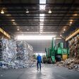 Ein Arbeiter aus der Ferne in einer großen Fabrikshalle, an den Seiten Müll für Recycling.
