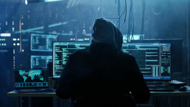 Eine Person in dunklem Kapuzensweater, nur von hinten sichtbar, sitzt an mehreren Computerbildschirmen, auf denen Datensätze zu sehen sind, in dunkler, gefährlich anmutender Umgebung