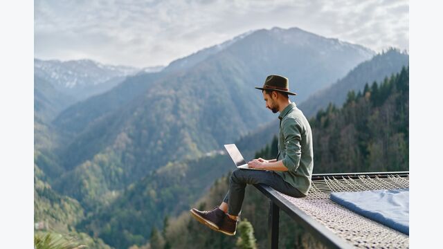 junger mann mit hut arbeitet am laptop auf kante eines trampolins sitzend, bergpanorama um ihn