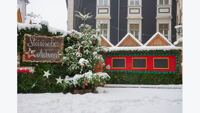 Ein menschenleerer steirischer Adventmarkt in Graz, mit Ständen und einem Weihnachtsbaum im Schnee