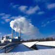 Schneebedeckte Solarpanele vor rauchender Fabrik unter blauem Himmel