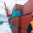 Ein Mensch in oranger Warnweste und orangem Schutzhelm steht bei hohem Wellengang auf einem Containerschiff, Rückenansicht.