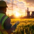Zwei Techniker in Warnwesten und Schutzhelmen stehen in einiger Entfernung zu einem Biomassekraftwerk in Betrieb, davor ein großes Feld.