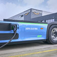 Der Rumpf eines blauen Elektro-Trucks (Modell eActros 600 von Mercedes) beim Tanken, im Hintergrund ein Amazon-Logistikgebäude.