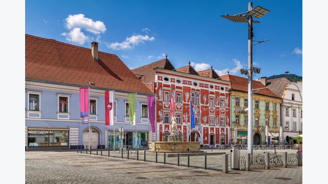 Der Hauptplatz in Leoben, Steiermark, mit bunten Häusern, Fahnen und Brunnen unter blauem Himmel.