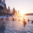 Eine ruhige Winterlandschaft mit Menschen bei traditionellem finnischen Saunaerlebnis in einen dampfenden See bei Sonnenuntergang, umgeben von schneebedeckten Bäumen.