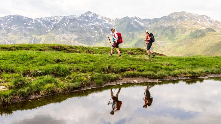 Zwei Frauen wandern bei schönem Wetter in den Alpen nahe einem kleinen Gewässer.