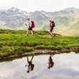 Zwei Frauen wandern bei schönem Wetter in den Alpen nahe einem kleinen Gewässer.