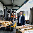 Alexander Winter, Obmann der Wiener Spediteure, steht unter dem dem Dachvorsprung eines Logistikbetriebs. Daneben sitzt ein Mann in oranger Warnweste in einem mit Holzpaletten beladenen Gabelstapler.