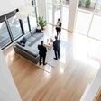 Immobilienmaklerin zeigt zwei Männern im Anzug ein modernes Wohnzimmer mit großer Fensterfront, Ansicht von oben.