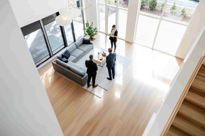 Immobilienmaklerin zeigt zwei Männern im Anzug ein modernes Wohnzimmer mit großer Fensterfront, Ansicht von oben.
