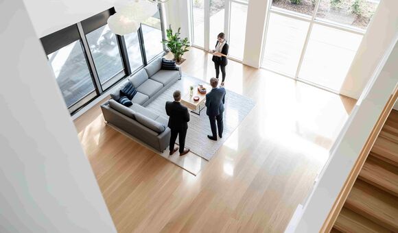Immobilienmaklerin zeigt zwei Männern im Anzug ein modernes Wohnzimmer mit großer Fensterfront, Ansicht von oben.