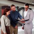 Ein Mann und eine Frau zwischen 30 und 40 lassen sich von einem Makler ein großes, modernes, leeres Haus zeigen und studieren gemeinsam die Exposé-Mappe.