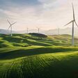 Mehrere Windräder unter blauem Himmel stehen auf einer hügeligen grünen Wiese, entfernt im Hintergrund Berge. KI-generiert