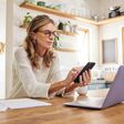 Eine reifere Frau sitzt an einem Laptop in einem Privathaushalt und blickt aufs Handy, neben ihr Papiere, die auf Buchhaltung, Steuererklärung oder Pensionsvorsorge schließen lassen.