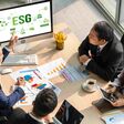 Luftansicht auf drei Männer und eine Frau in formeller Kleidung, die in einem Büro an einem Tisch sitzen und ein Meeting abhalten, vor sich Geschäftsberichte und ein Bildschirm, auf dem groß "ESG" steht.
