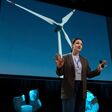 Eine junge Frau hält in einem dunklen Raum einen Vortrag, hinter ihr eine Windturbine abgebildet auf einem riesigen Bildschirm.