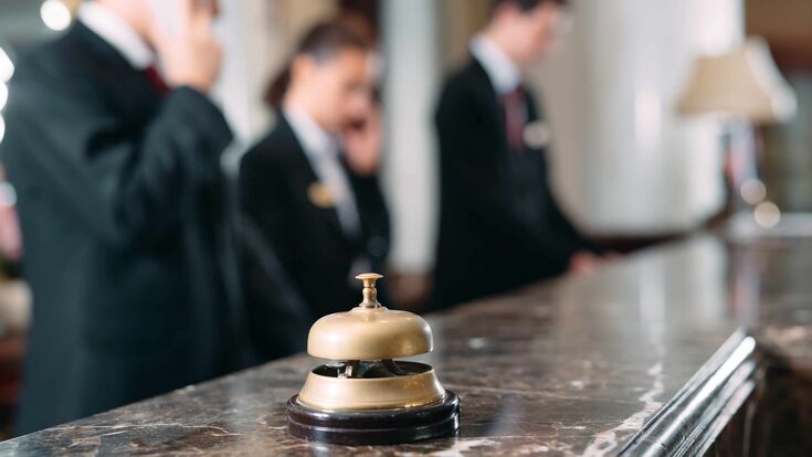 © adobe stock/davit85 Drei Rezeptionisten in einem Hotel bei der Arbeit, zwei davon am Telefon, alle verschwommen im Hintergrund. Im Vordergrund eine Glocke auf dem Tresen.
