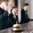 Drei Rezeptionisten in einem Hotel bei der Arbeit, zwei davon am Telefon, alle verschwommen im Hintergrund. Im Vordergrund eine Glocke auf dem Tresen.