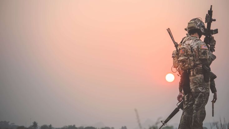 © Adobe Stock/Chinnachote Zwei Soldaten von schräg hinten, die bei fahl scheinender Sonne in voller Militärmontur schwere Waffen halten.