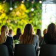 Rückenansicht auf mehrere sitzende Personen bei einer Konferenz, bei Podium sind verschwommen große grüne Pflanzen zu sehen.
