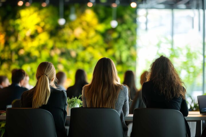 Rückenansicht auf mehrere sitzende Personen bei einer Konferenz, bei Podium sind verschwommen große grüne Pflanzen zu sehen.