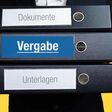 Person im Anzug trägt einen Stapel mit 3 Aktenordnern, der mittlere trägt die Aufschrift "Vergabe".