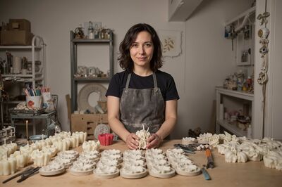 © adobe Stock/S u n n y B u n n y KI-generiertes Bild einer lächelnden jungen Frau an einem großen Werktisch mit vielen selbstgemachten Kerzen vor sich.