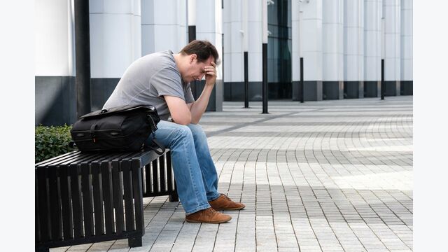 Ein niedergeschlagener Mann mittleren Alters sitzt auf einer Bank vor einem Gebäude, neben sich eine Tasche, den Kopf in die Hand gestürzt.