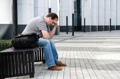 Ein niedergeschlagener Mann mittleren Alters sitzt auf einer Bank vor einem Gebäude, neben sich eine Tasche, den Kopf in die Hand gestürzt.