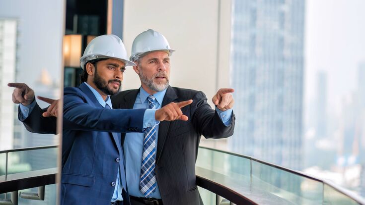 © Adobe Stock/Tongpool Ein junger und ein älterer Manager auf dem Balkon eines Hochhauses, mit Sicherheitshelmen, blicken und zeigen auf einen Punkt in der Distanz.