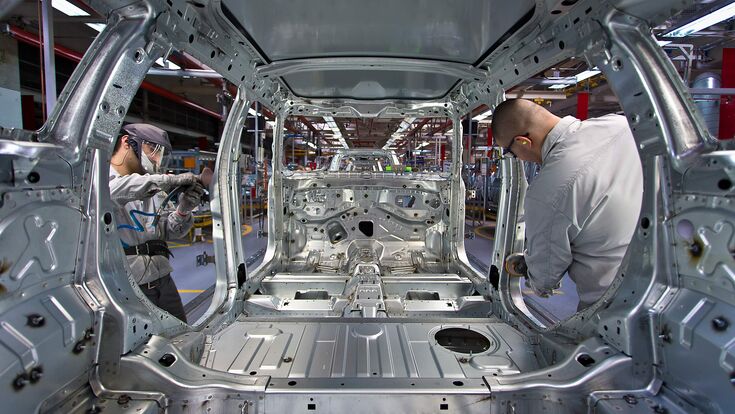 © dieindustrie.at/Mathias Kniepeiss Arbeiter werken an einem chromfarbenen Rohbau eines Autos in Industrieumgebung.