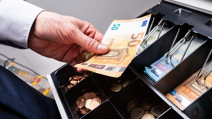 Eine Hand gibt einen 50-Euro-Schein in eine professionelle, große Geldkasse voll mit scheinen und Münzen.