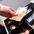 Eine Hand gibt einen 50-Euro-Schein in eine professionelle, große Geldkasse voll mit scheinen und Münzen.