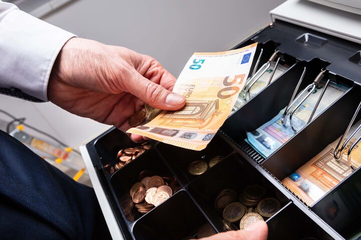 Eine Hand gibt einen 50-Euro-Schein in eine professionelle, große Geldkasse voll mit scheinen und Münzen.