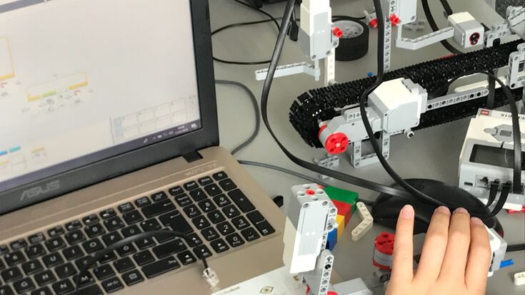 © RoboManiac Kinderhand auf einer Computermaus neben Laptop, auf dem Mindstorm zu sehen ist.