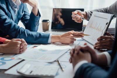 © Adobe Stock/NanSan Business Meeting mehrerer Personen an einem Tisch, im Mittelpunkt ein Klemmbrett mit einem Chart.