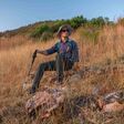 Wanderer mit Hut, Rucksack, Sonnenbrille und nordic walking stock sitzt auf einem trocken begrasten Hügel und blickt in die Ferne.