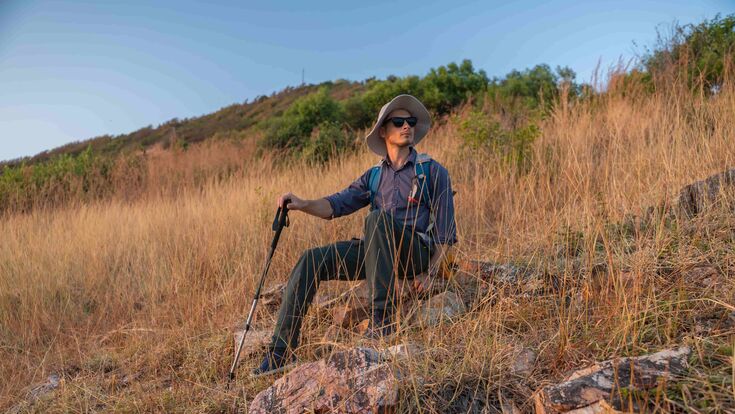 © Adobe stock/APchanel Wanderer mit Hut, Rucksack, Sonnenbrille und nordic walking stock sitzt auf einem trocken begrasten Hügel und blickt in die Ferne.