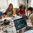 Mehrere Kinder, zwischen 10 und 12 Jahren, an einem großen Tisch mit Laptops und kleinen Roboterteilen.