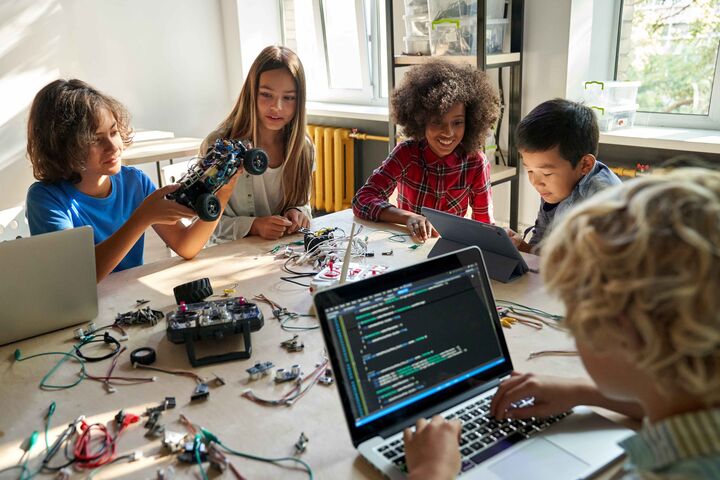 Mehrere Kinder, zwischen 10 und 12 Jahren, an einem großen Tisch mit Laptops und kleinen Roboterteilen.