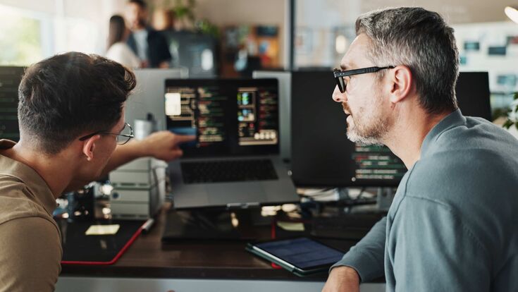 Ein junger und ein etwas älterer Mann bei der Programmierarbeit am Computer im Büro: Der jüngere erklärt dem älteren etwas und deutet auf ein laufendes Programm am Laptop
