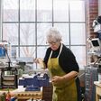 Eine alte, weißhaarige Frau spannt in einer Werkstatt ein Stück Holz ein.