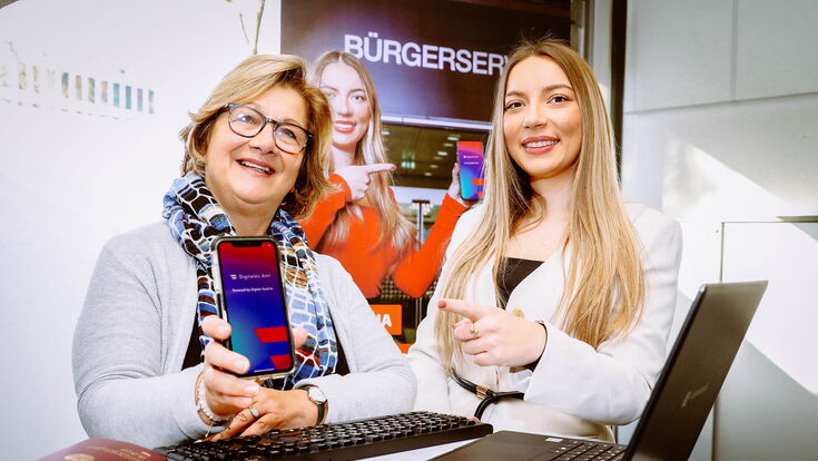 Zwei Frauen an einem Infostand mit offenem Laptop bewerben  „Digital Überall“-Workshops im Digital-Angebot der Stadt Leoben.