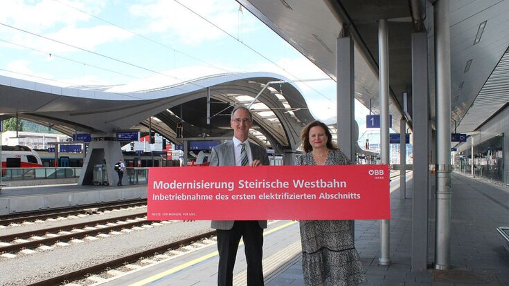 ÖBB-Geschäftsführer Ewald Jantscher und LRin Claudia Holzer halten auf einem Bahnhof ein rotes Schild in die Höhe, mit Aufschrift: "Modernisierung Steirische Westbahn"