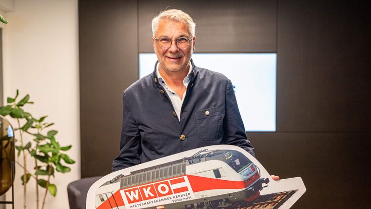 Rudolf Bredschneider, Obmann der WK-Bezirksstelle Völkermarkt, mit einem großen Schild, das einen Zug zeigt.